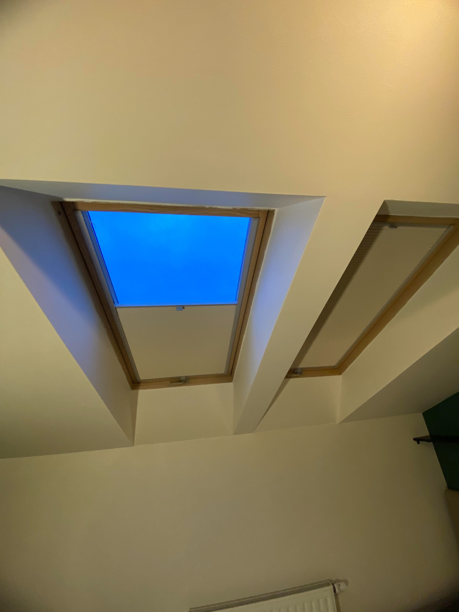 Dwa okna dachowe w skosie sufitu, jedno z widokiem na błękitne niebo, drugie z zamontowaną plisą w kolorze kremowym.