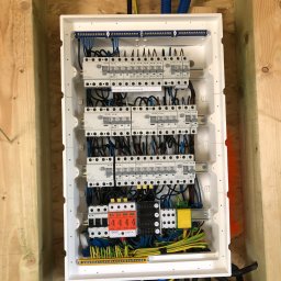 Tomasz Ossowski ELTOM - Rozdzielnica elektryczna w białej obudowie, zamontowana w konstrukcji drewnianej, widoczne bezpieczniki, przewody w kolorach niebieskim, żółto-zielonym i czarnym, oraz peszle ochronne w kolorze...