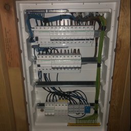 Tomasz Ossowski ELTOM - Rozdzielnia elektryczna z zabezpieczeniami różnicowoprądowymi i nadprądowymi marki Schneider, z widocznymi przewodami w kolorach niebieskim, czarnym i zielono-żółtym, zamontowana na drewnianej...