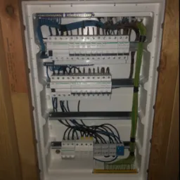 Rozdzielnia elektryczna z zabezpieczeniami różnicowoprądowymi i nadprądowymi marki Schneider, z widocznymi przewodami w kolorach niebieskim, czarnym i zielono-żółtym, zamontowana na drewnianej...