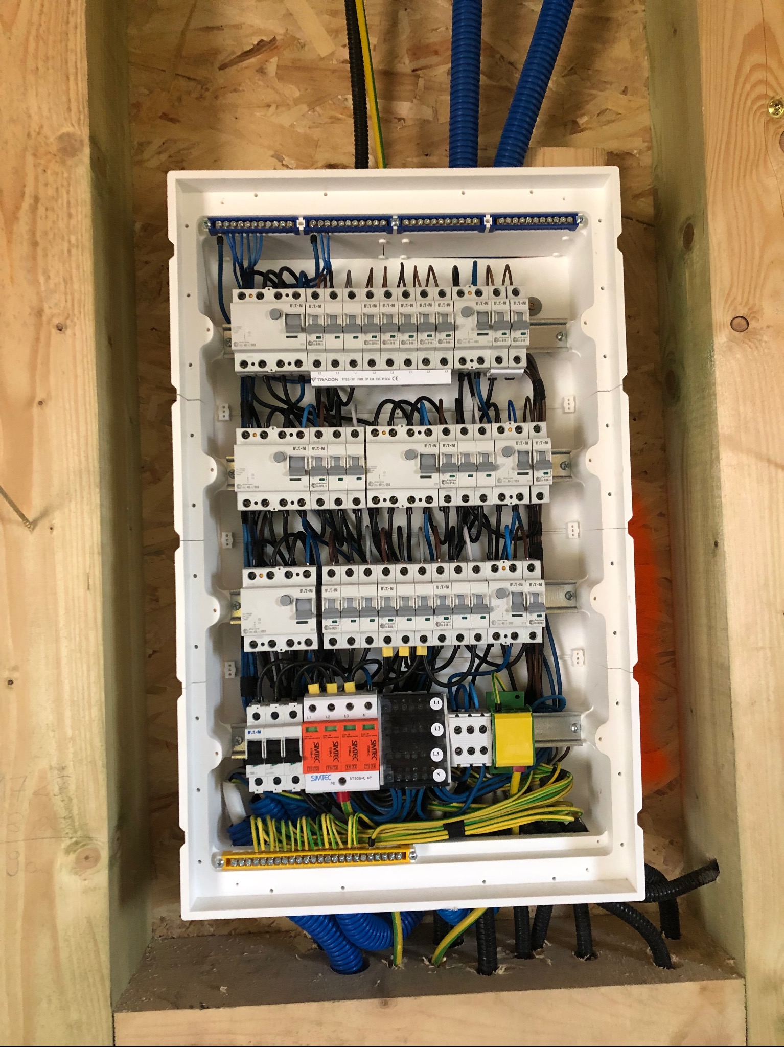 Rozdzielnica elektryczna w obudowie zamontowana w drewnianej konstrukcji ściany, widoczne liczne bezpieczniki, przewody w różnych kolorach, listwy zaciskowe i peszle ochronne.