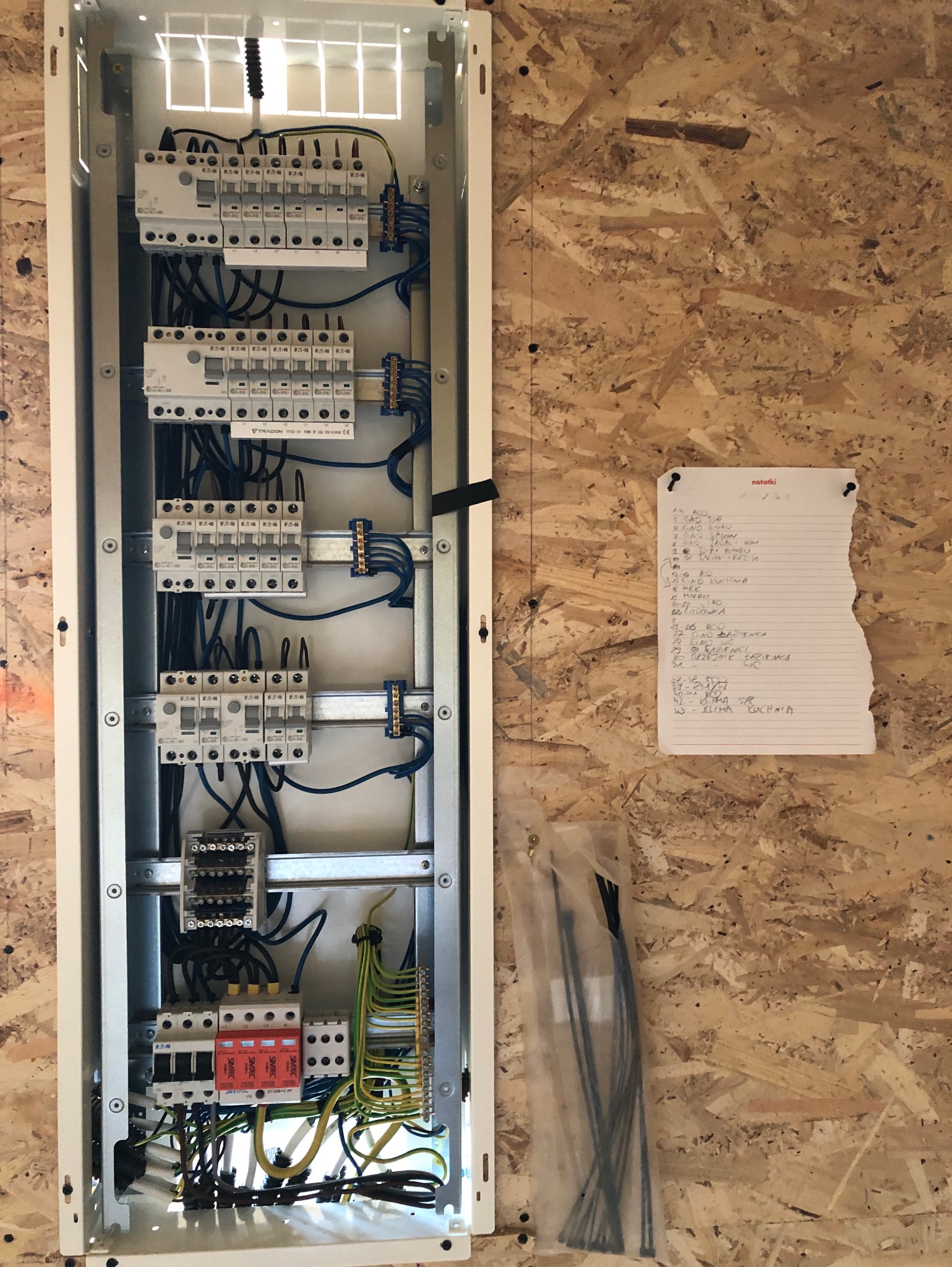 Rozdzielnica elektryczna w trakcie montażu, widoczne zabezpieczenia, przewody i listwa zaciskowa, obok notatki z planem instalacji na ścianie z płyty OSB.