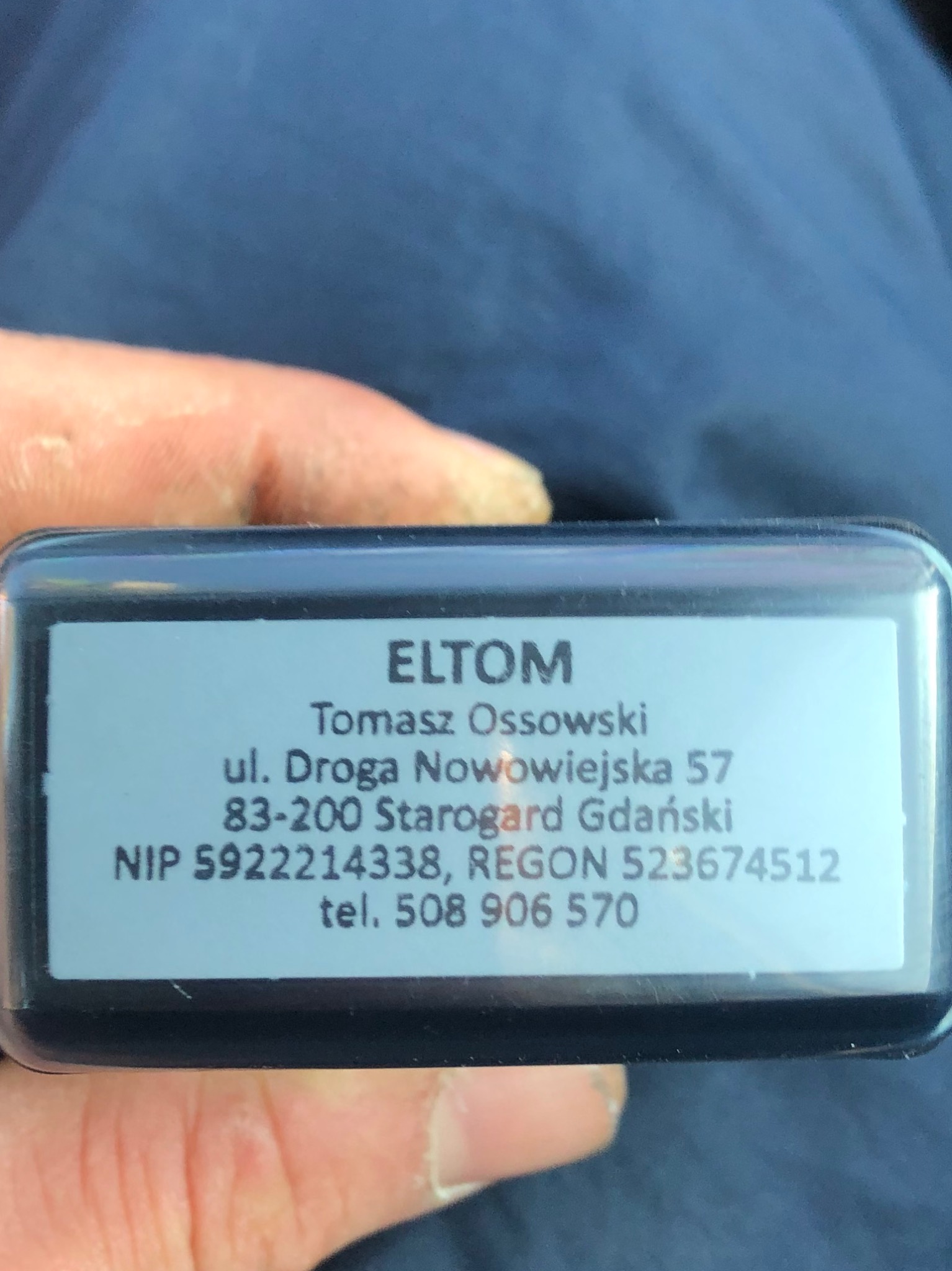 Fragment wizytówki firmy ELTOM Tomasz Ossowski z adresem i numerem telefonu, trzymanej w dłoni na tle niebieskiego materiału.