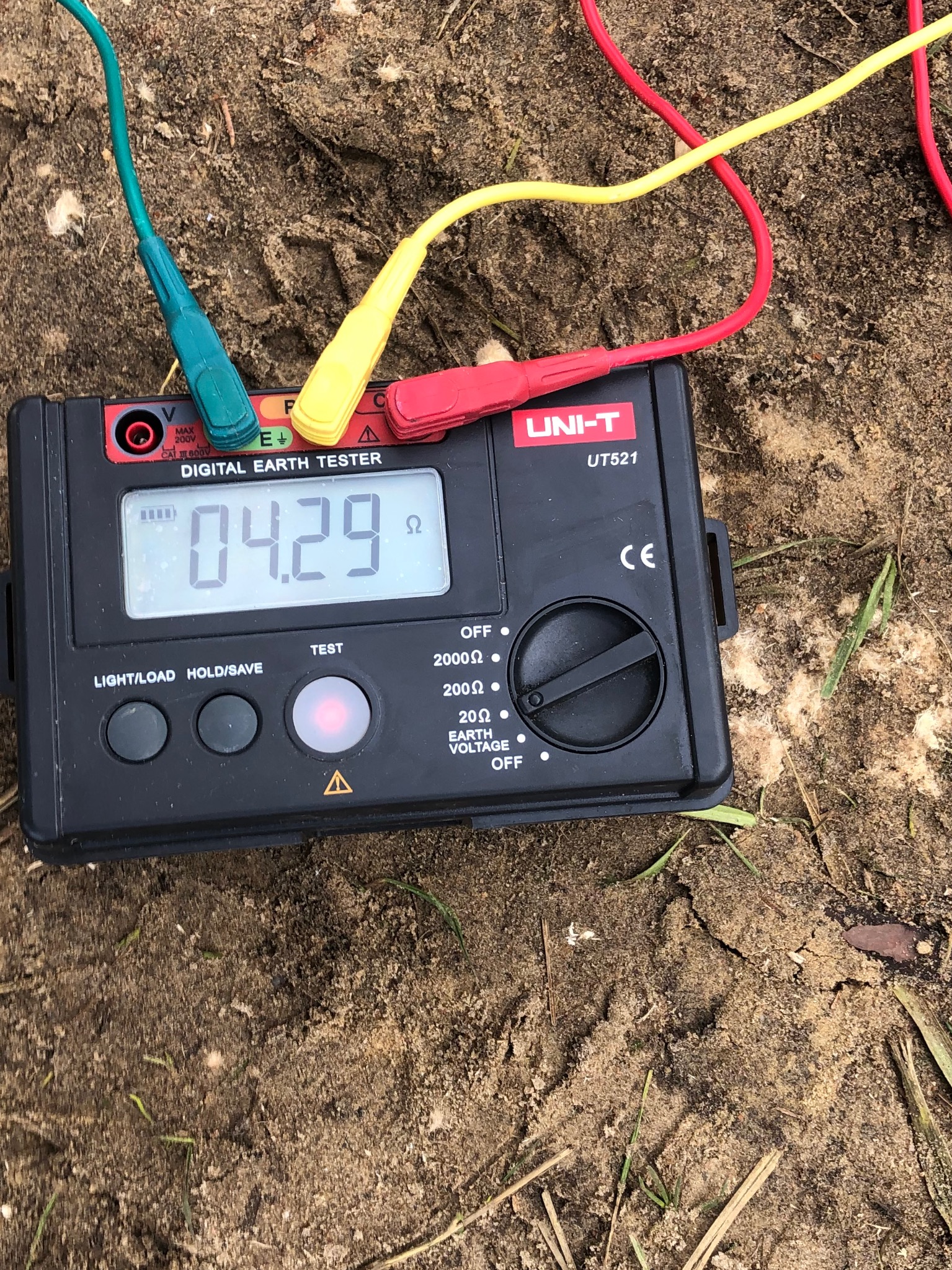 Miernik cyfrowy UNI-T UT521 do pomiaru uziemienia z podłączonymi przewodami pomiarowymi w kolorach zielonym, żółtym i czerwonym, leżący na ziemi, wskazujący wartość 04.29 Ω.