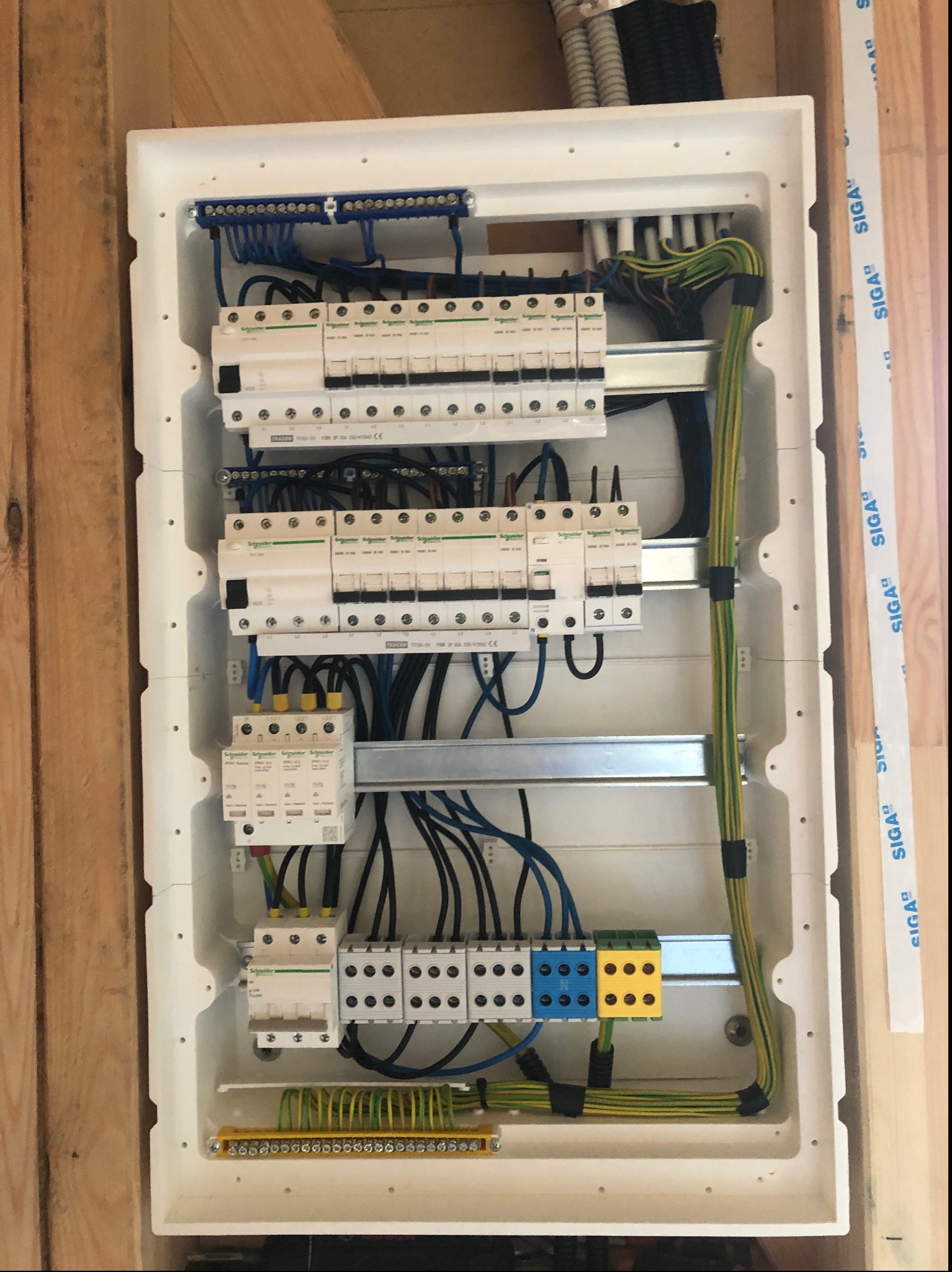 Rozdzielnica elektryczna Schneider w trakcie montażu, widoczne zabezpieczenia, listwy zaciskowe, okablowanie w kolorach niebieskim, czarnym i żółto-zielonym, zamontowana w ścianie z drewnianych desek.