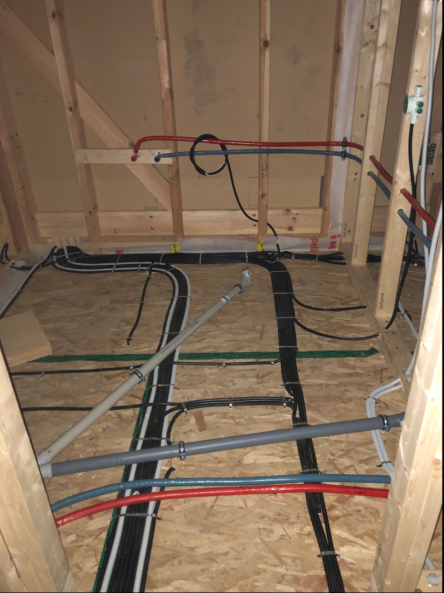 Wnętrze poddasza w trakcie instalacji: widoczne liczne kable elektryczne i rury, przymocowane do drewnianej konstrukcji dachu i podłogi z płyty OSB za pomocą metalowych uchwytów. Część przewodów...