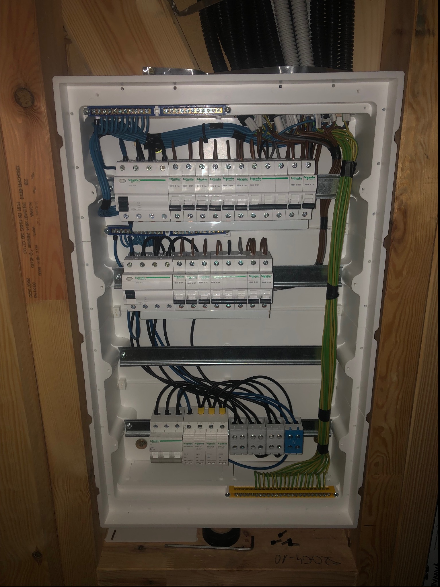 Rozdzielnia elektryczna z zabezpieczeniami różnicowoprądowymi i nadprądowymi marki Schneider, z widocznymi przewodami w kolorach niebieskim, czarnym i zielono-żółtym, zamontowana na drewnianej...