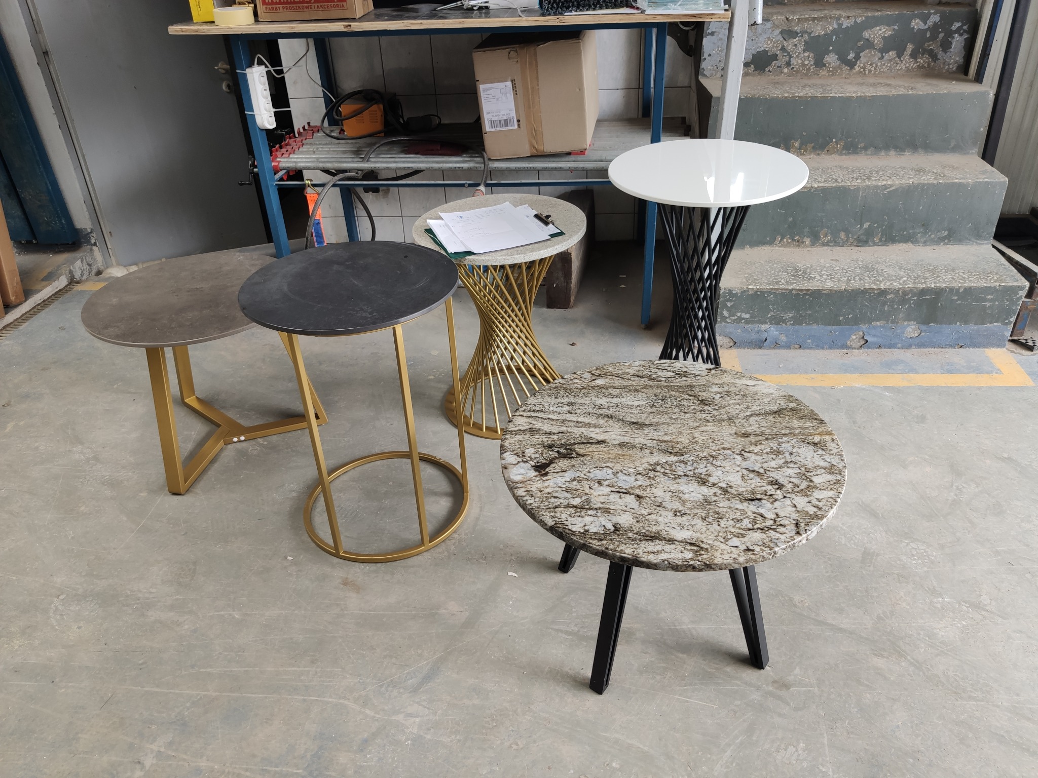 Cztery małe stoliki z różnymi blatami (kamienny, konglomerat, biały) i podstawami (złota, czarna), ustawione w warsztacie lub magazynie.
