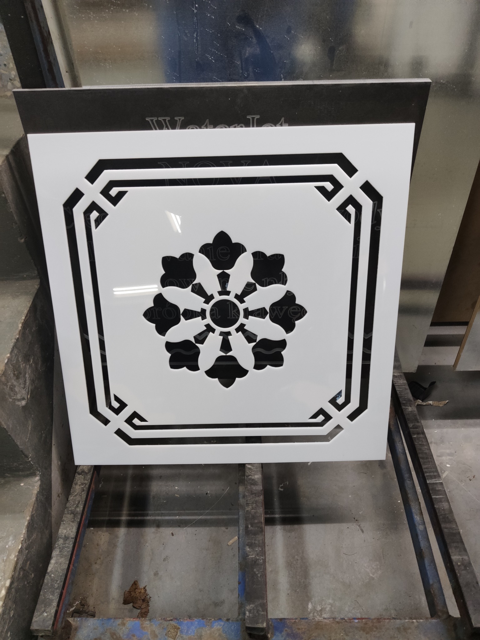 Biały, kwadratowy panel dekoracyjny z wyciętym wzorem kwiatowym i geometrycznym, ustawiony na metalowej konstrukcji.