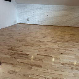 PrimeFinish Sebastian Krupa - Jasne, świeżo polakierowane deski podłogowe z widocznymi refleksami światła, ułożone w przestronnym pomieszczeniu z białą ceglaną ścianą i skośnym sufitem.