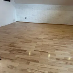 Jasne, świeżo polakierowane deski podłogowe z widocznymi refleksami światła, ułożone w przestronnym pomieszczeniu z białą ceglaną ścianą i skośnym sufitem.