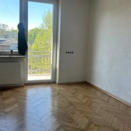 PrimeFinish Sebastian Krupa - Jasny parkiet w jodełkę w pokoju z oknem balkonowym i grzejnikiem. Widoczne listwy przypodłogowe i gniazdka elektryczne. Dobre oświetlenie naturalne.
