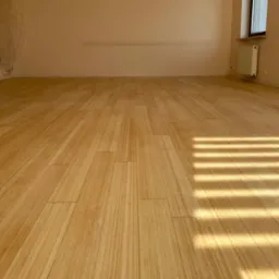 Jasna podłoga z paneli, z widocznym wzorem słojów, oświetlona promieniami słońca wpadającymi przez okno. Ściany beżowe, widoczny grzejnik.