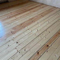 PrimeFinish Sebastian Krupa - Jasna, odnowiona podłoga z desek w pokoju z grzejnikiem i surowymi ścianami. Widoczne naturalne usłojenie drewna po renowacji.
