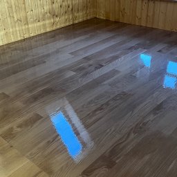 PrimeFinish Sebastian Krupa - Błyszcząca, odnowiona podłoga z paneli w pokoju na poddaszu, odbijająca światło z okna dachowego i okna ściennego. Ściany i sufit wykończone boazerią.