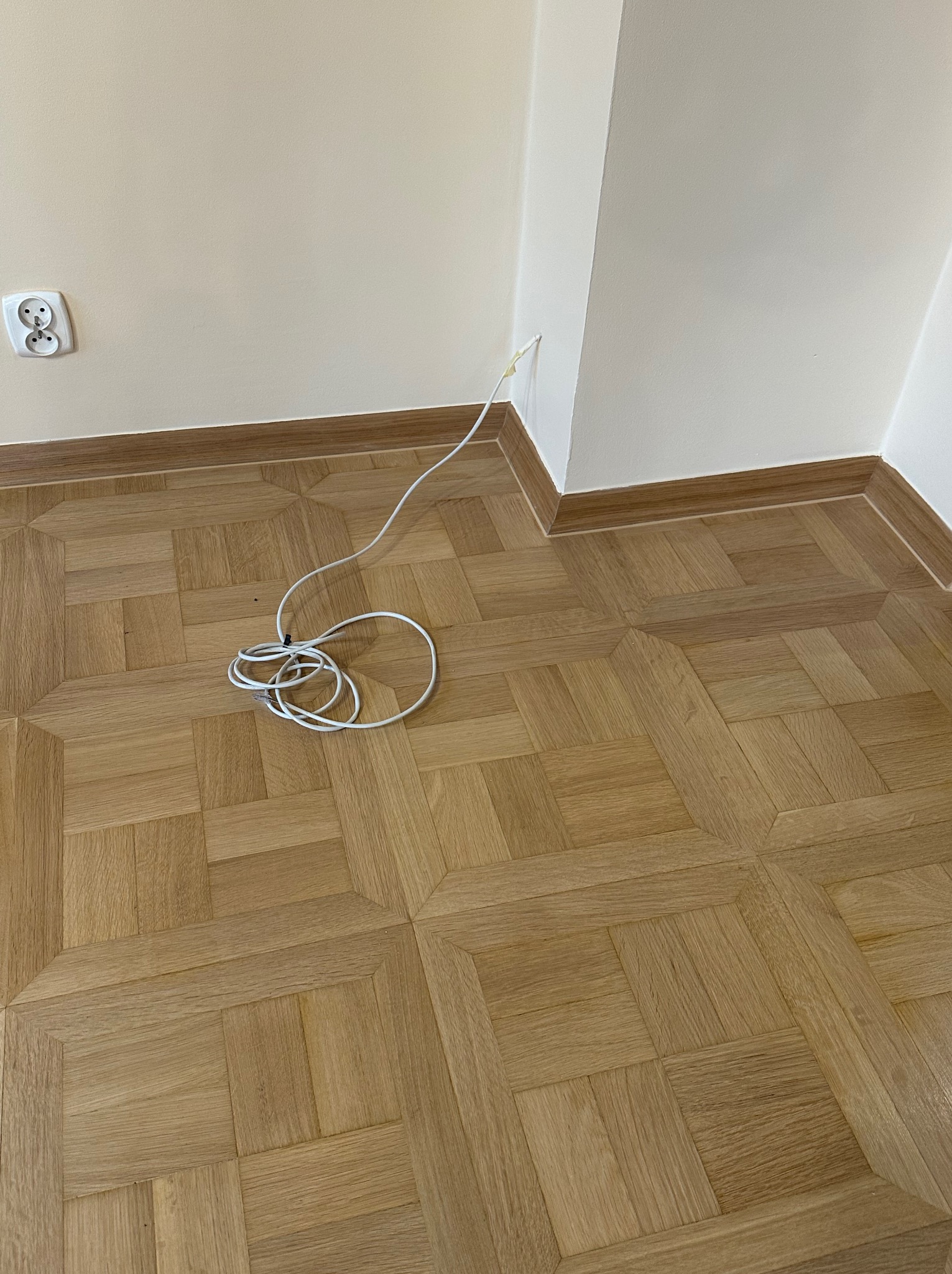 Parkiet ułożony w kwadraty z widocznym kablem wystającym ze ściany i zwiniętym na podłodze, przy listwie przypodłogowej.
