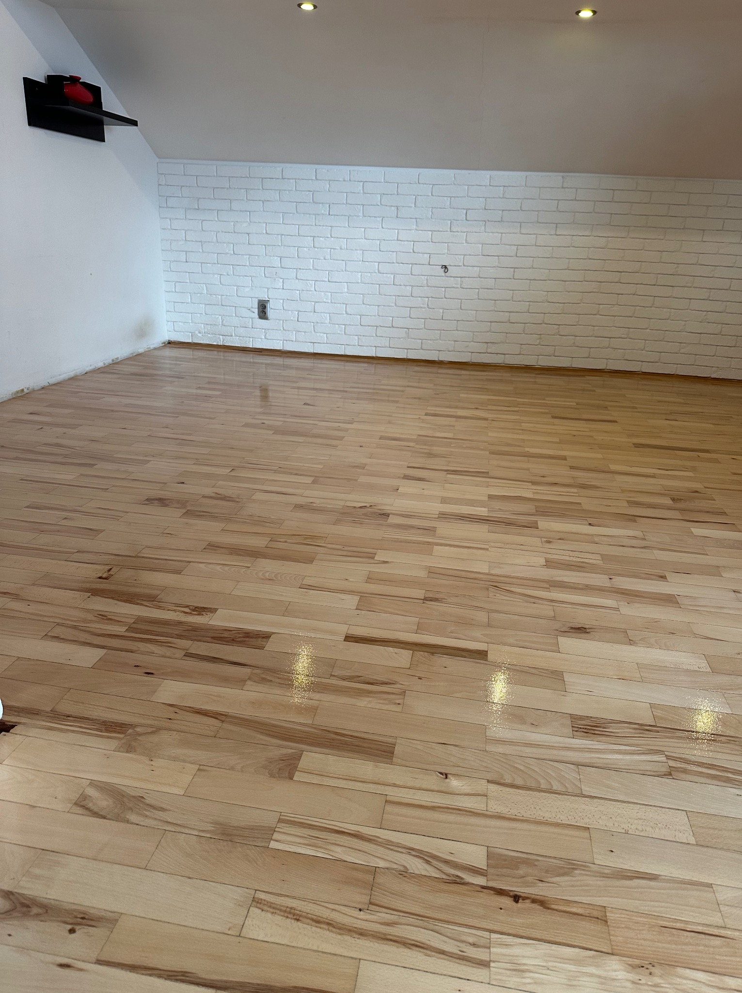 Jasne, świeżo polakierowane deski podłogowe z widocznymi refleksami światła, ułożone w przestronnym pomieszczeniu z białą ceglaną ścianą i skośnym sufitem.