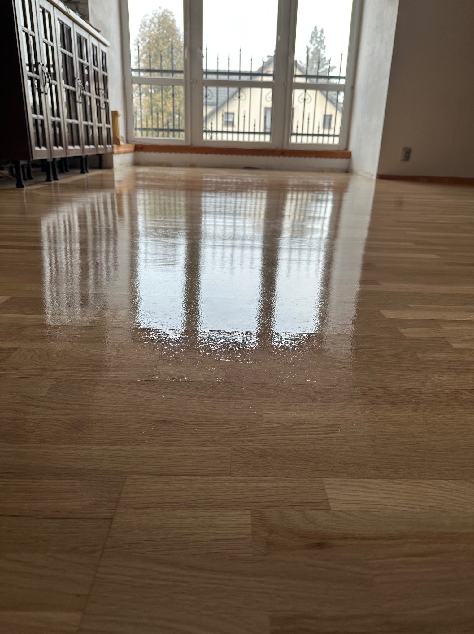 Świeżo polakierowana podłoga z paneli drewnianych, odbijająca okno z widokiem na dom i drzewa, w tle ciemny mebel.