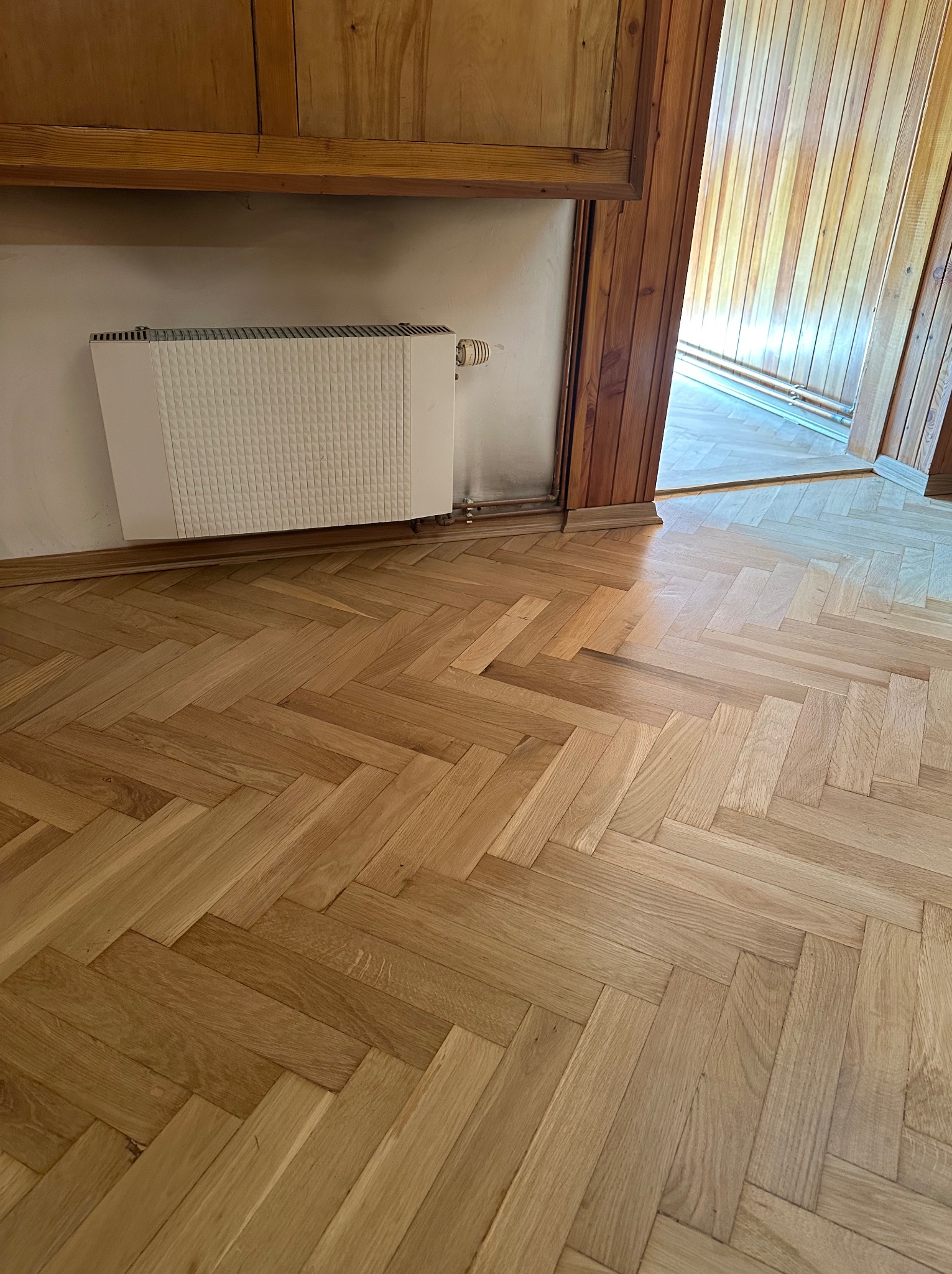 Parkiet ułożony w jodełkę, jasne drewno, widoczny fragment grzejnika i listwy przypodłogowe. Renowacja podłogi w mieszkaniu.