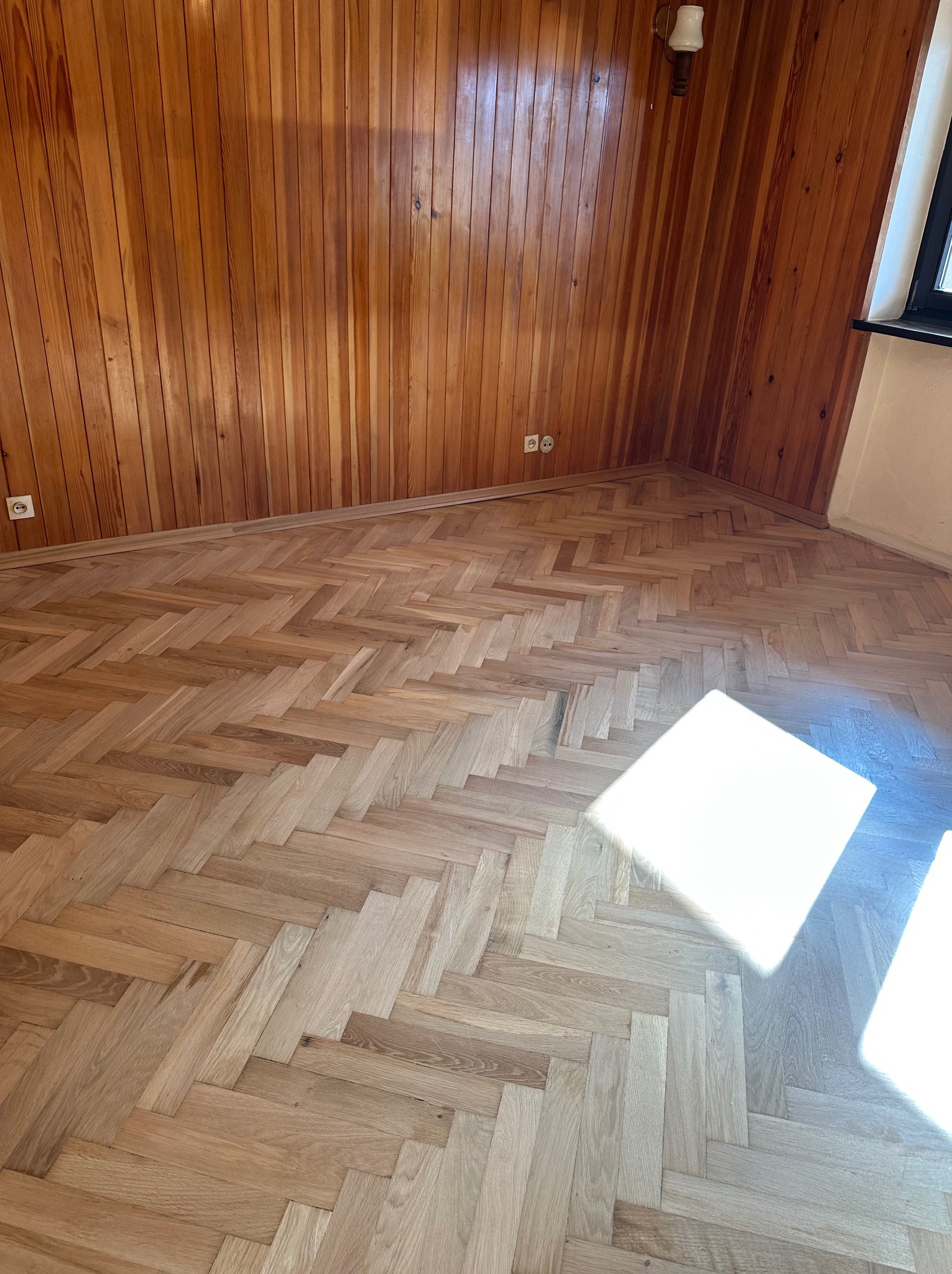 Jasny, parkiet ułożony w jodełkę, po renowacji, z widocznymi listwami przypodłogowymi i drewnianą boazerią na ścianach. W rogu pokoju widoczny fragment okna.
