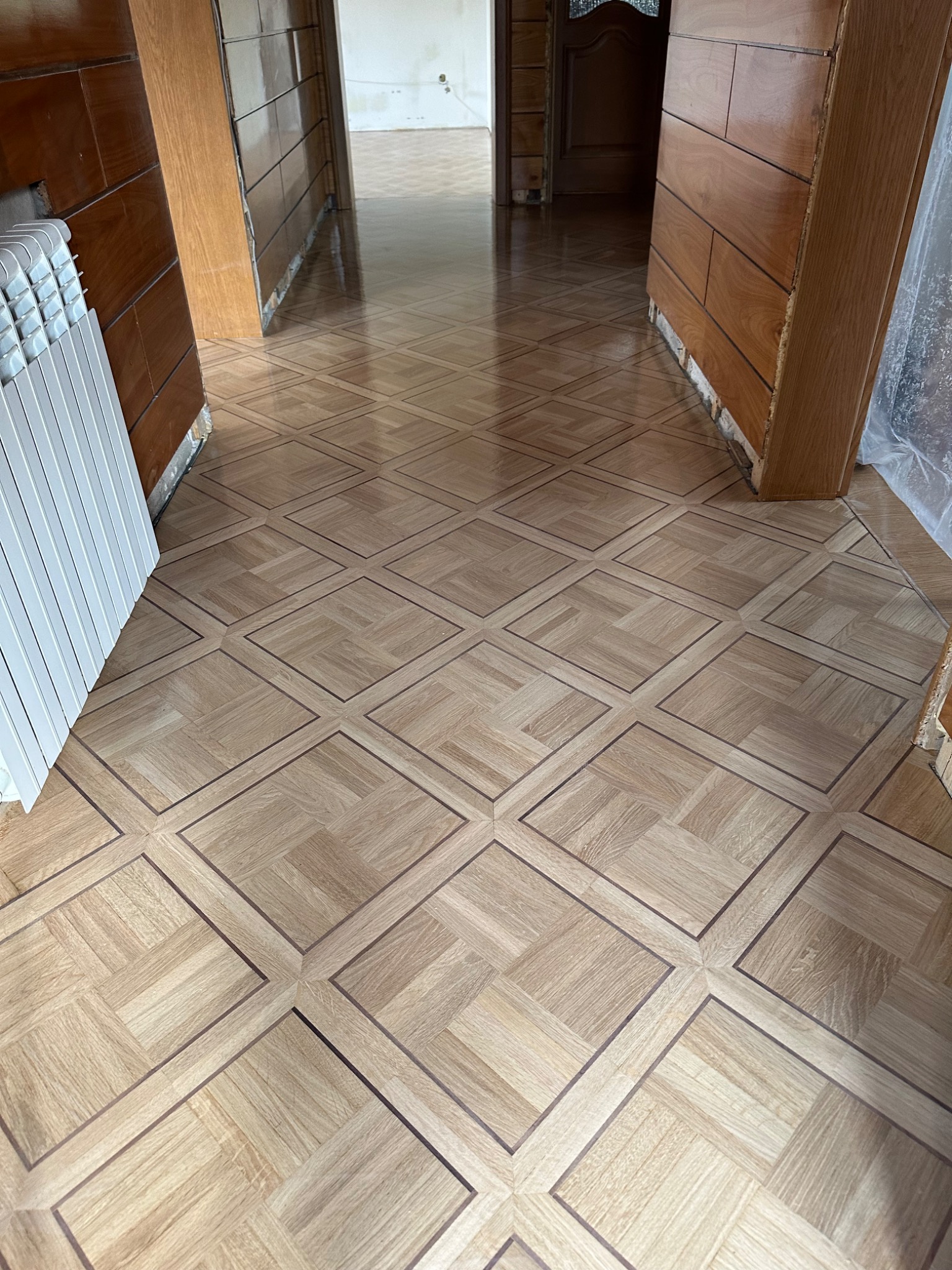 Odnowiona podłoga z parkietu ułożonego we wzór kwadratów, widok z perspektywy korytarza, z fragmentem grzejnika po lewej stronie. Widoczne ślady prac wykończeniowych.