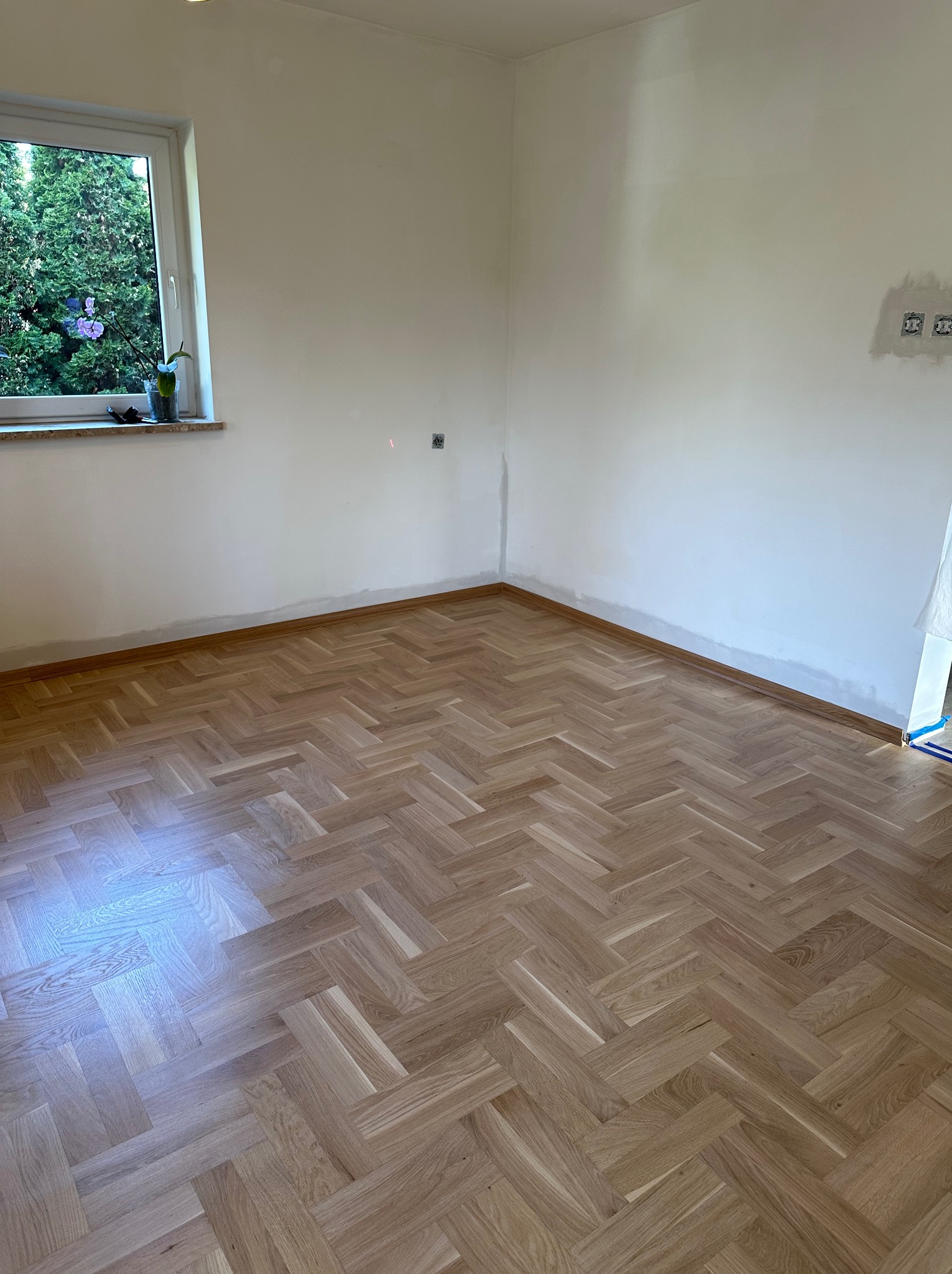 Jasny parkiet ułożony w jodełkę francuską w pustym pokoju z oknem. Świeżo pomalowane ściany, brak listew przypodłogowych. Widoczny laser na ścianie.