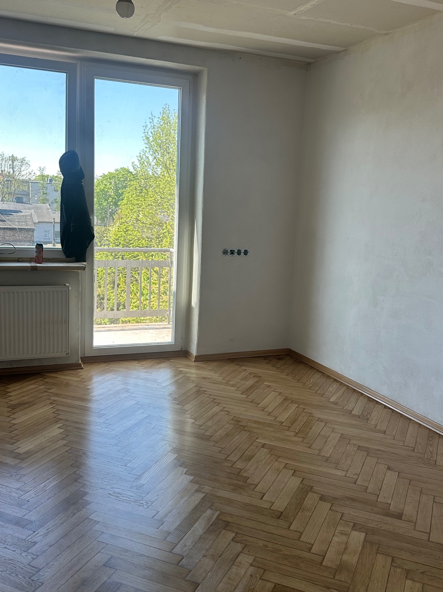 Jasny parkiet w jodełkę w pokoju z oknem balkonowym i grzejnikiem. Widoczne listwy przypodłogowe i gniazdka elektryczne. Dobre oświetlenie naturalne.