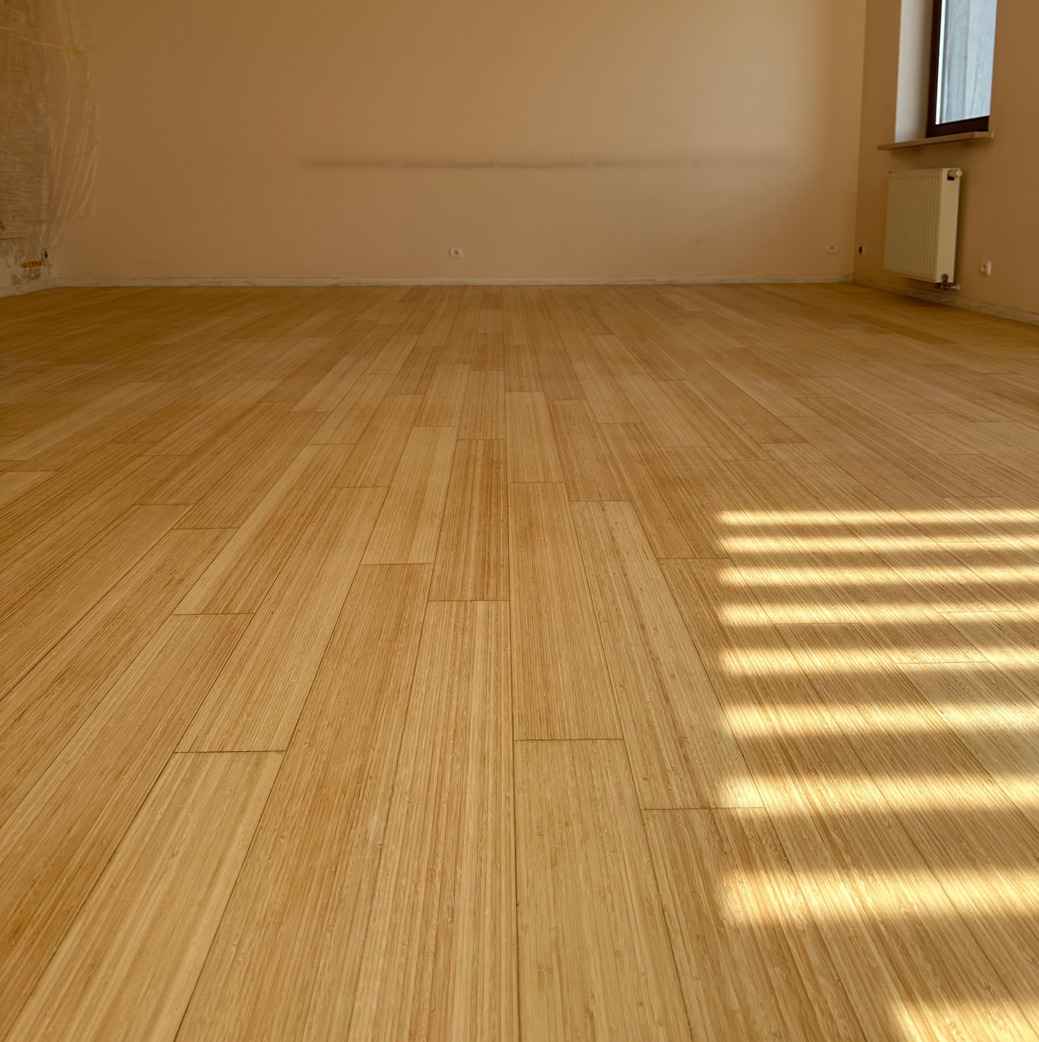 Jasna podłoga z paneli, z widocznym wzorem słojów, oświetlona promieniami słońca wpadającymi przez okno. Ściany beżowe, widoczny grzejnik.