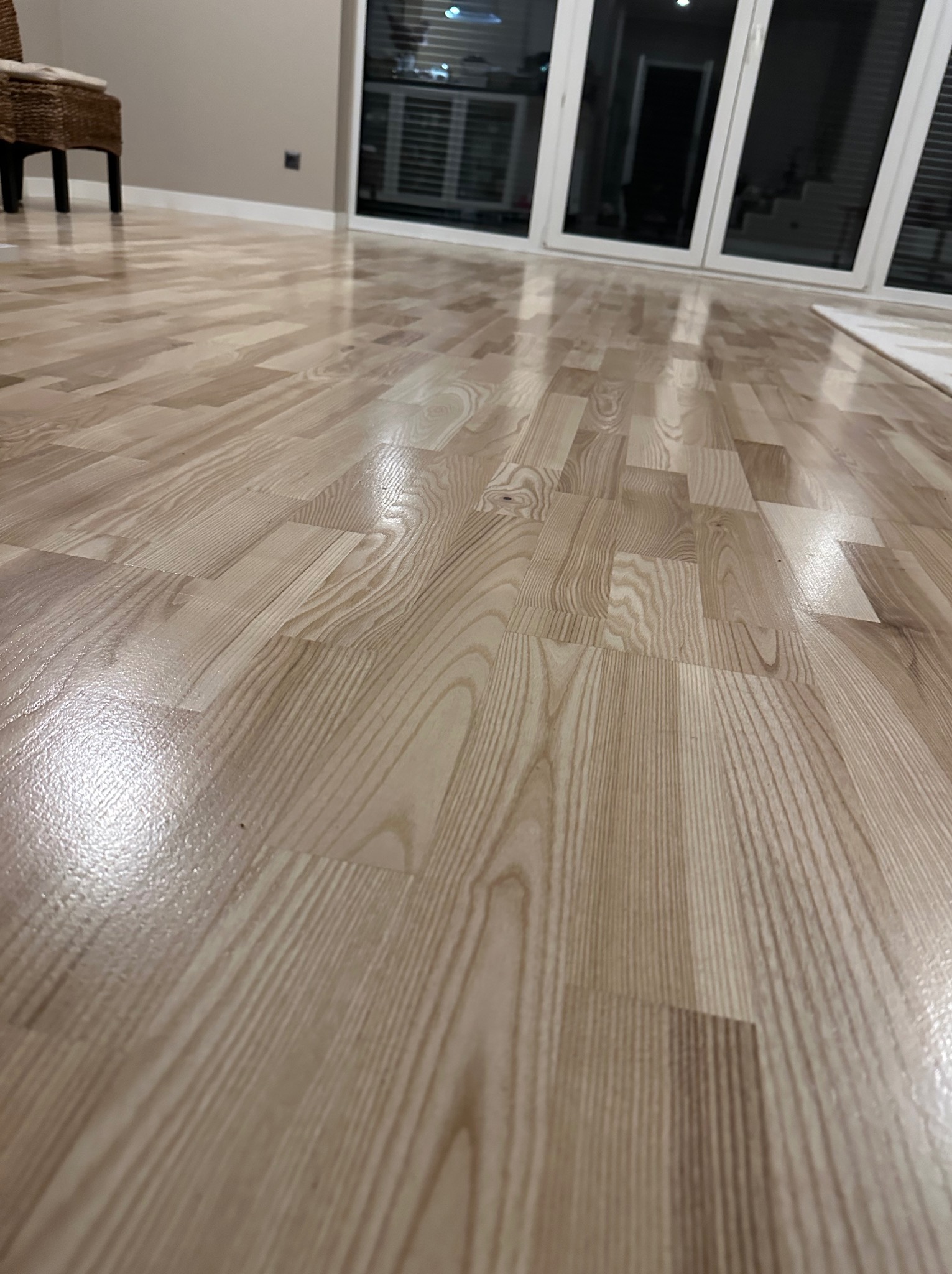 Jasne panele podłogowe ułożone w jodełkę, z widocznym odbiciem światła, w tle przesuwne drzwi balkonowe i fragment wiklinowego fotela.