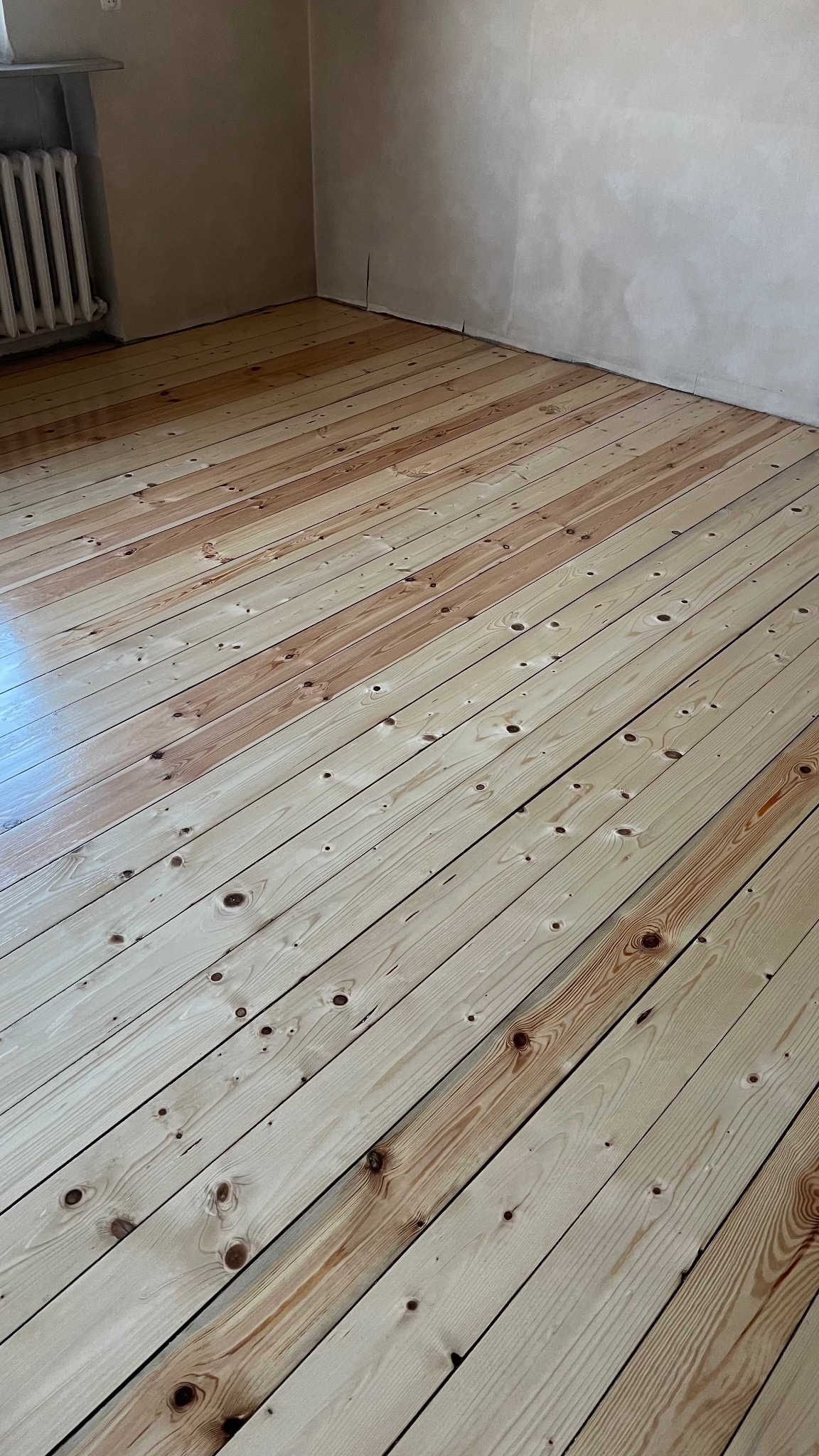 Jasna, odnowiona podłoga z desek w pokoju z grzejnikiem i surowymi ścianami. Widoczne naturalne usłojenie drewna po renowacji.