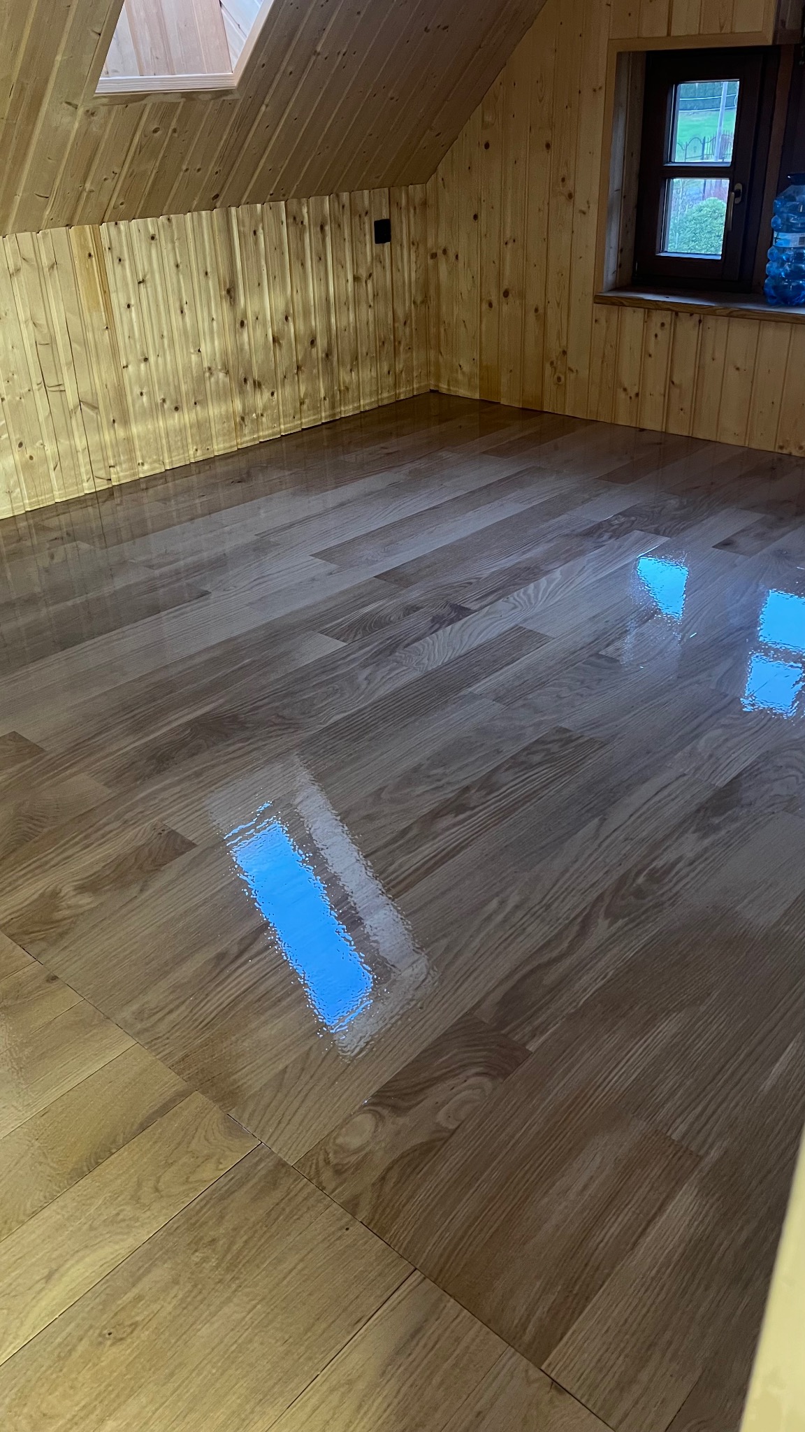 Błyszcząca, odnowiona podłoga z paneli w pokoju na poddaszu, odbijająca światło z okna dachowego i okna ściennego. Ściany i sufit wykończone boazerią.