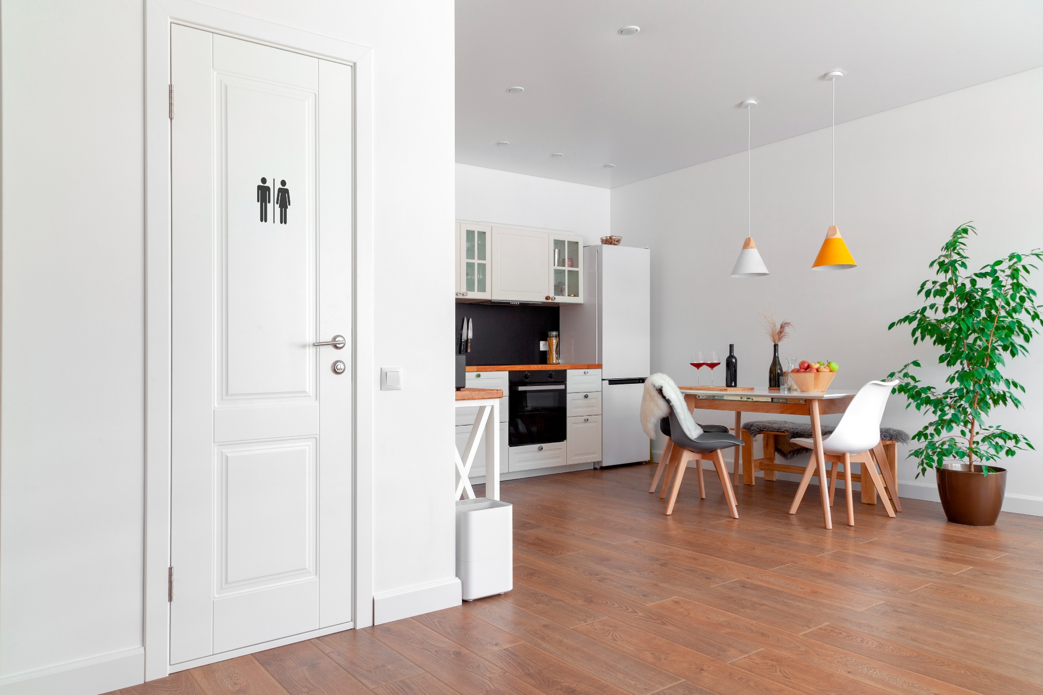 Wnętrze mieszkania w stylu skandynawskim: drewniana podłoga, białe ściany, drzwi z symbolem toalety, kuchnia w tle, stół z owocami i winem, roślina doniczkowa.