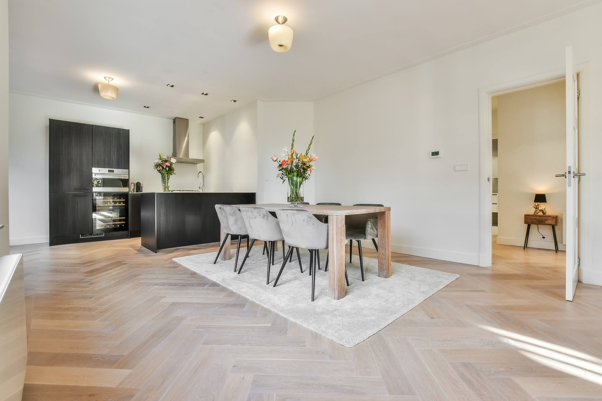 Jasny parkiet w jodełkę w przestronnym wnętrzu z otwartą kuchnią, ciemnymi szafkami, drewnianym stołem z szarymi krzesłami i kwiatami w wazonie.
