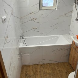 Usługi budowlane - Wykończona łazienka z białą wanną obudowaną płytkami imitującymi marmur, drewnopodobną podłogą i oknem. Widoczny bojler i umywalka z szafką.