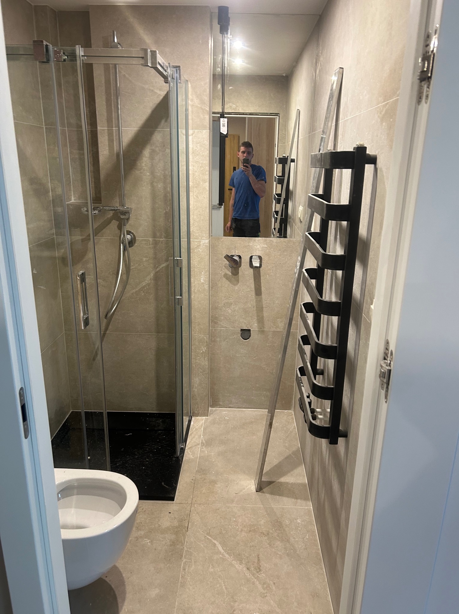 Wykończona łazienka z kabiną prysznicową, toaletą i grzejnikiem drabinkowym. Beżowe płytki na ścianach i podłodze. W lustrze odbija się osoba robiąca zdjęcie.