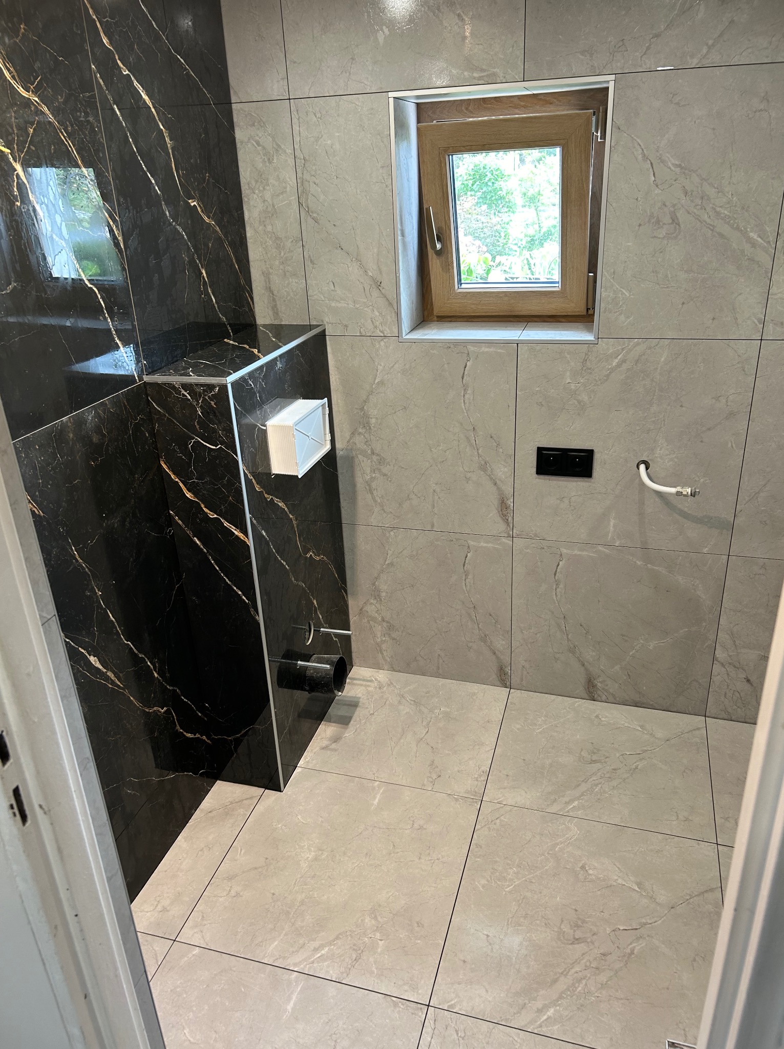 Nowoczesna łazienka z marmurowymi płytkami w odcieniach szarości i czerni. Widoczne okno, zabudowa toalety i fragment instalacji. Minimalistyczny design.
