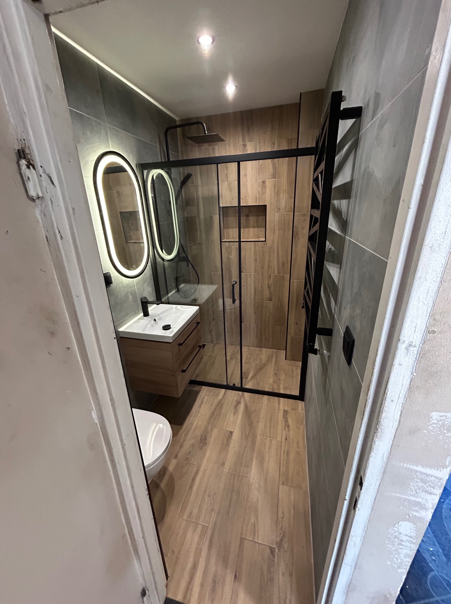 Nowoczesna łazienka po remoncie: kabina prysznicowa typu walk-in z czarnymi ramami, podświetlane lustra owalne nad drewnianą szafką z umywalką, płytki imitujące drewno i szarość.