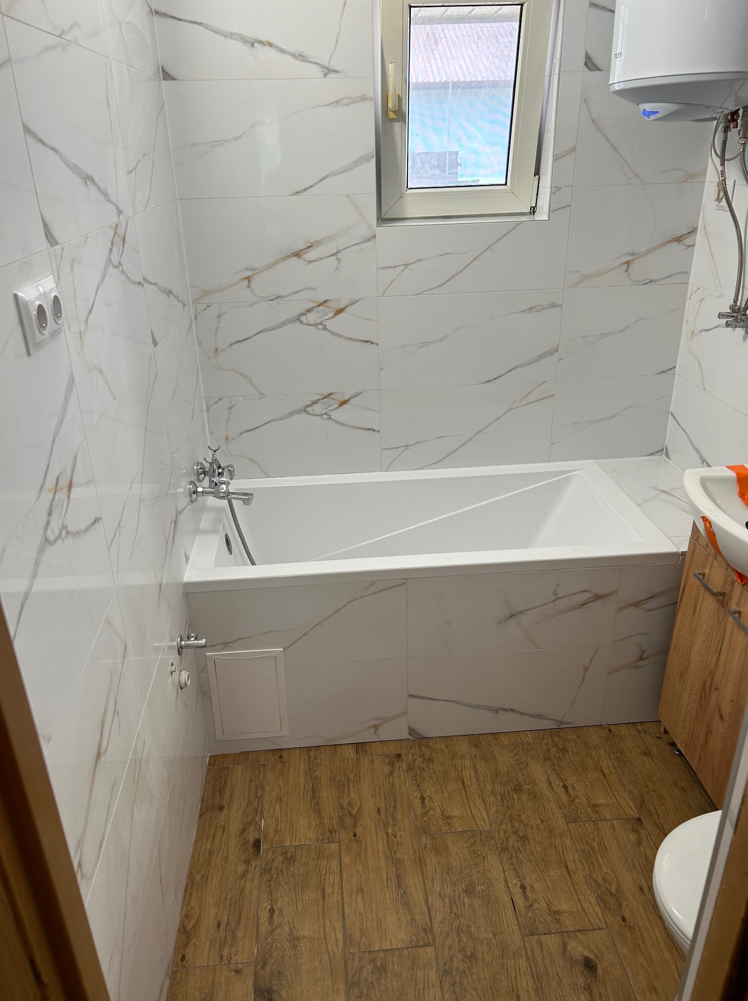 Wykończona łazienka z białą wanną obudowaną płytkami imitującymi marmur, drewnopodobną podłogą i oknem. Widoczny bojler i umywalka z szafką.