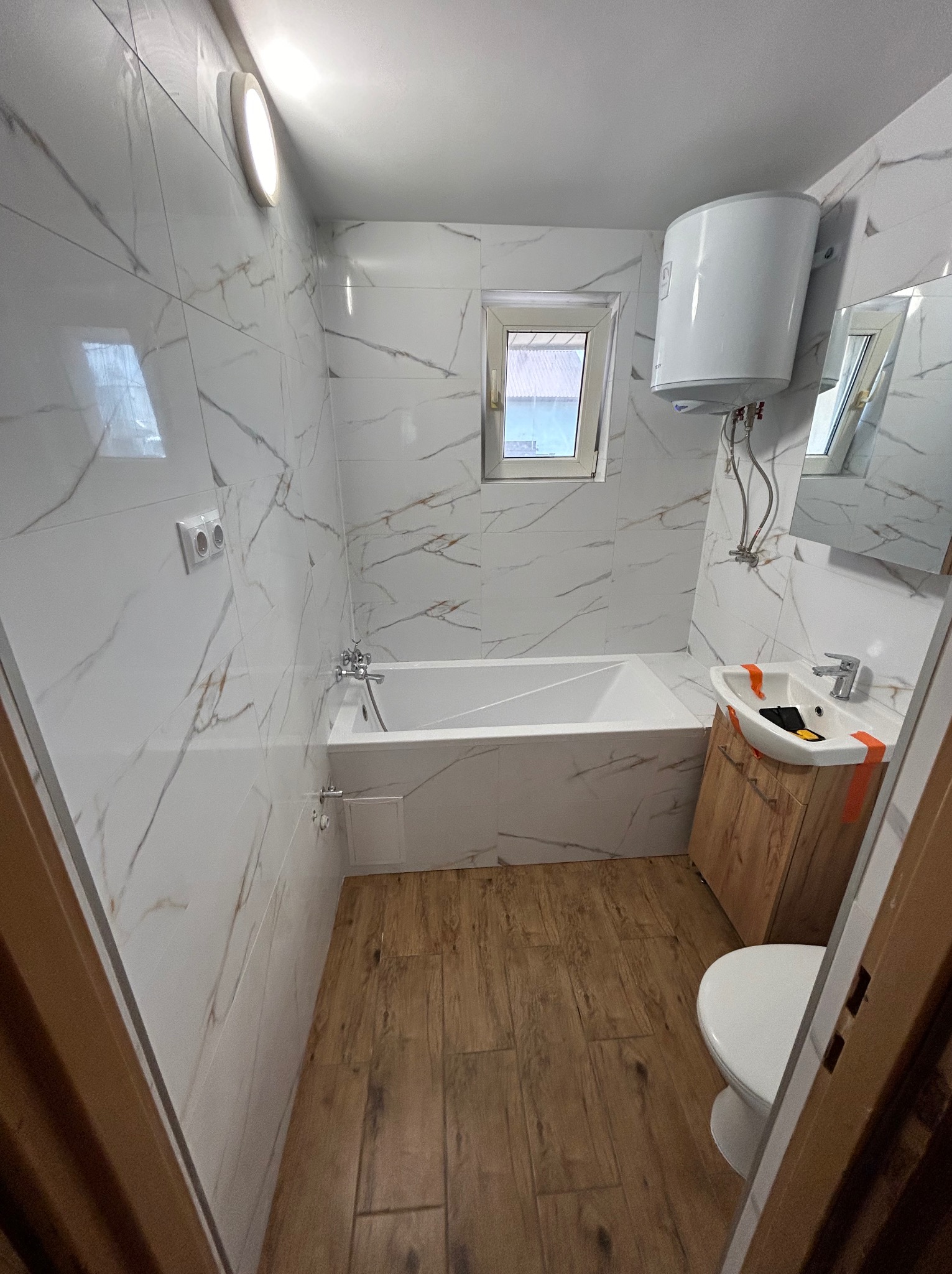 Wykończona łazienka z białą wanną, umywalką na szafce z drewnianym dekorem i toaletą. Ściany wyłożone białymi płytkami z szarym marmurkowym wzorem, drewnopodobna podłoga.