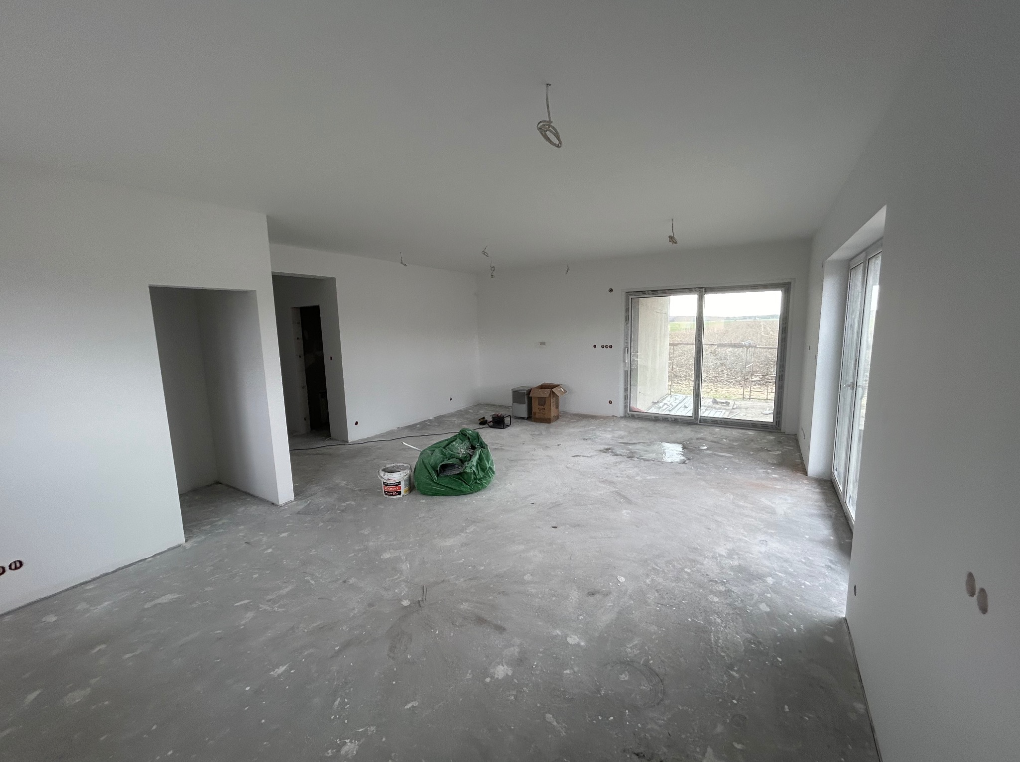 Wnętrze w trakcie wykończenia: białe ściany, surowa podłoga, okno balkonowe. Widoczne kable, puszki elektryczne, wiadro z farbą i worek z odpadami budowlanymi.
