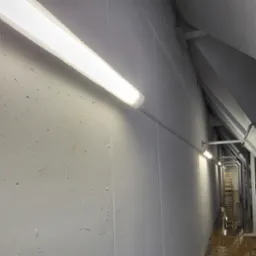 Długa lampa fluorescencyjna na szarej ścianie z widocznymi śladami po malowaniu. W tle widoczny korytarz z drabinką i oświetleniem punktowym.