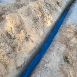 Wykop w piasku z niebieską folią zabezpieczającą, prawdopodobnie pod kable elektryczne. Widoczne grudki ziemi i drobne kamienie w wykopie.