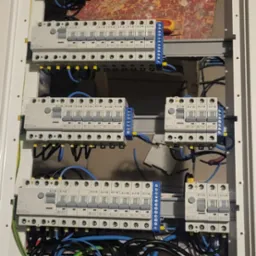 Rozdzielnia elektryczna w białej obudowie, widoczne zabezpieczenia różnicowoprądowe, bezpieczniki, listwy zaciskowe i okablowanie. Zasilanie w trakcie podłączania.