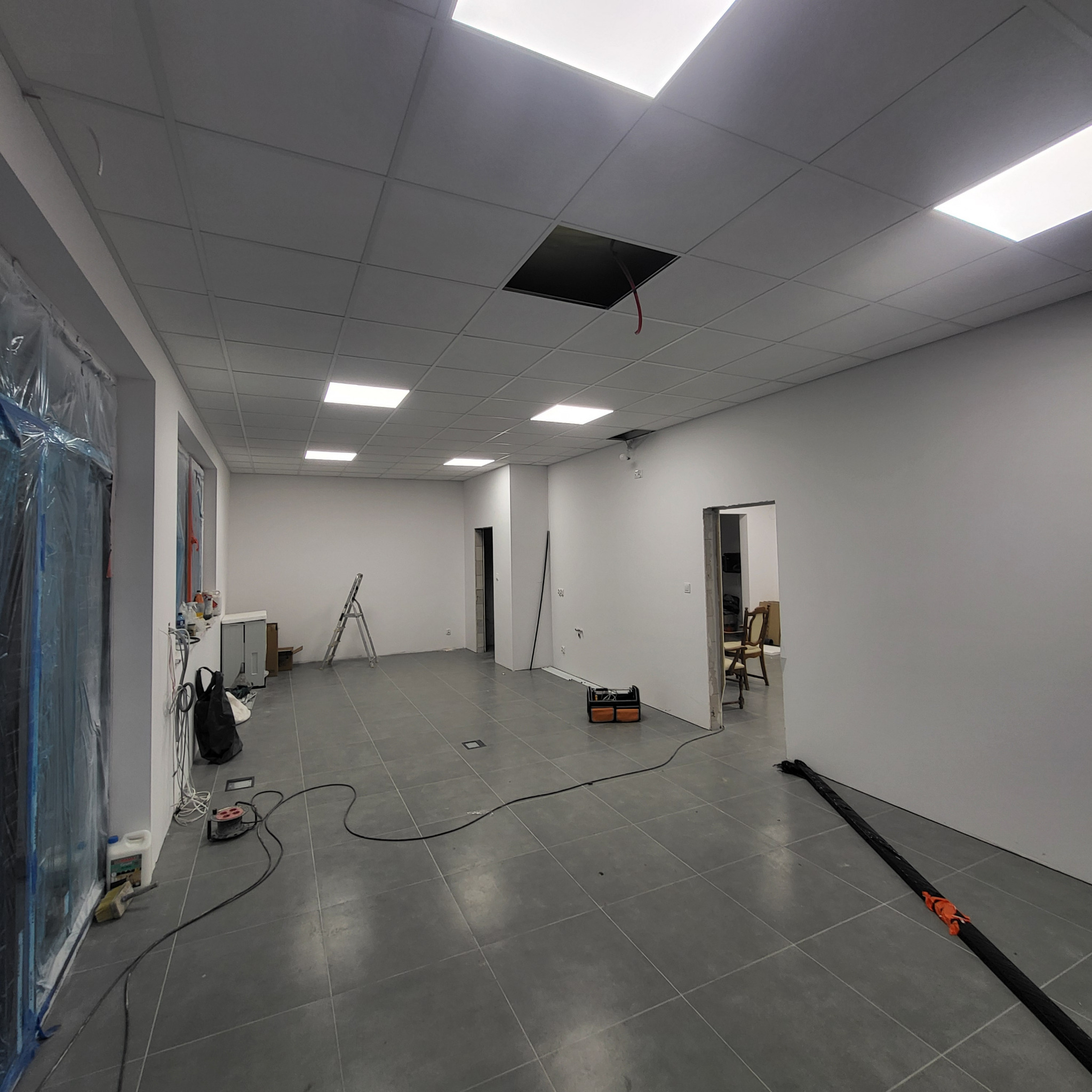 Wnętrze w trakcie wykańczania: szara podłoga, białe ściany, sufit podwieszany z oświetleniem. Widoczne narzędzia i kable wskazują na prace elektryczne.
