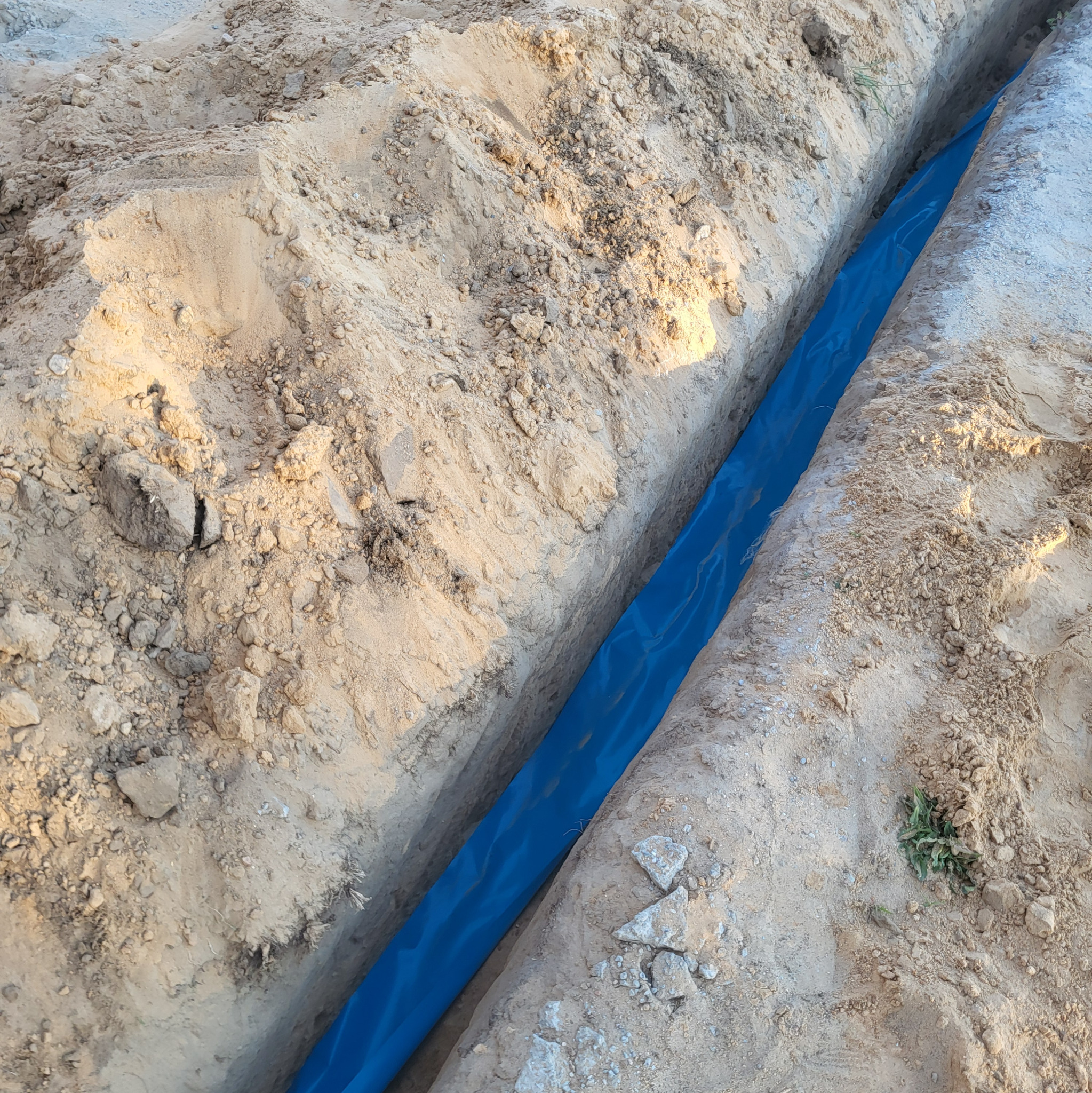 Wykop w piasku z niebieską folią zabezpieczającą, prawdopodobnie pod kable elektryczne. Widoczne grudki ziemi i drobne kamienie w wykopie.