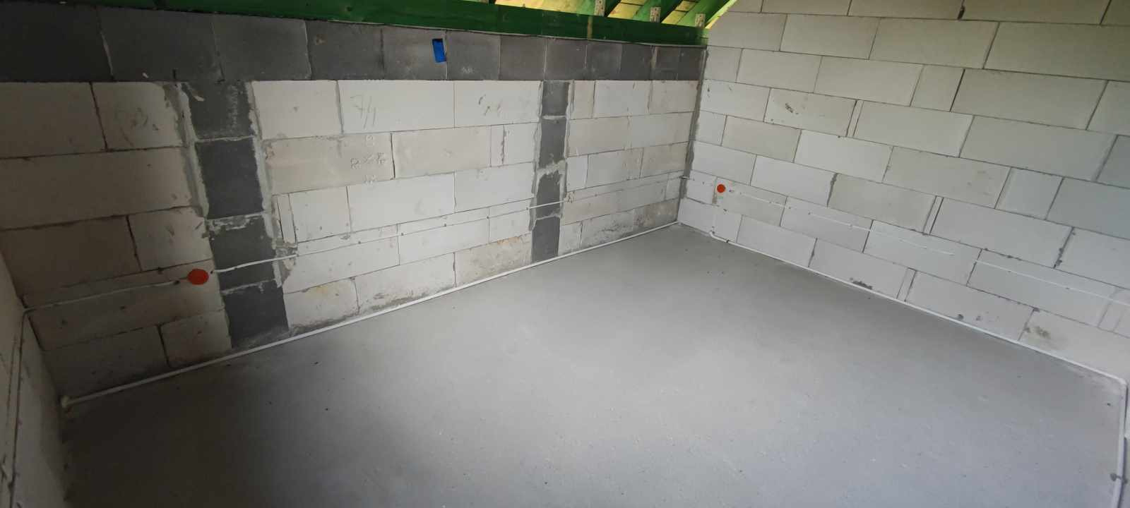 Surowy stan wnętrza: ściany z białych bloczków, częściowo zaszpachlowane, widoczne przewody elektryczne z pomarańczowymi puszkami, wylana szlichta podłogowa.