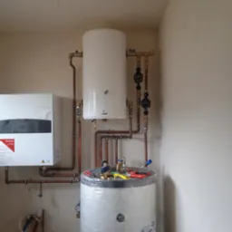 Biały bojler akumulacyjny owinięty folią, obok piec gazowy Ariston, widoczne miedziane rury, zawory oraz manometry, instalacja w trakcie montażu.