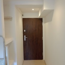 RM Produkcja - Korytarz po remoncie: świeżo pomalowane, białe ściany z brązowymi drzwiami wejściowymi. Widoczne ślady po instalacji na ścianie.