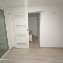 RM Produkcja - Białe, przesuwne drzwi z poziomymi przeszkleniami, zamontowane w jasnym wnętrzu. Widoczny fragment okna balkonowego i podłoga imitująca drewno.