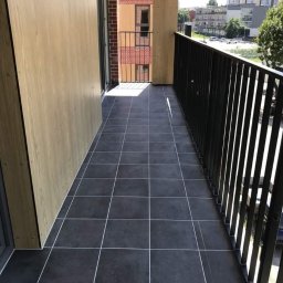 RM Produkcja - Wykończony balkon z ciemnymi, kwadratowymi płytkami i jasnymi fugami. Widok na zabudowę miejską. Nowoczesne balustrady i drewniane panele boczne.